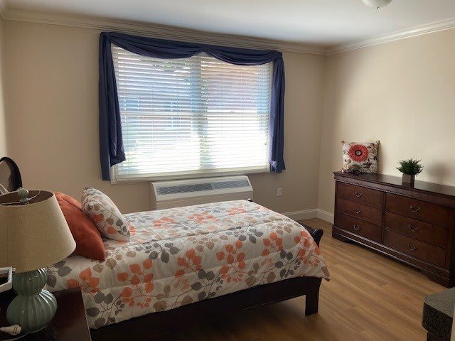A cozy bedroom with a single bed and floral bedding, featuring a large window with curtains and a wooden dresser.
