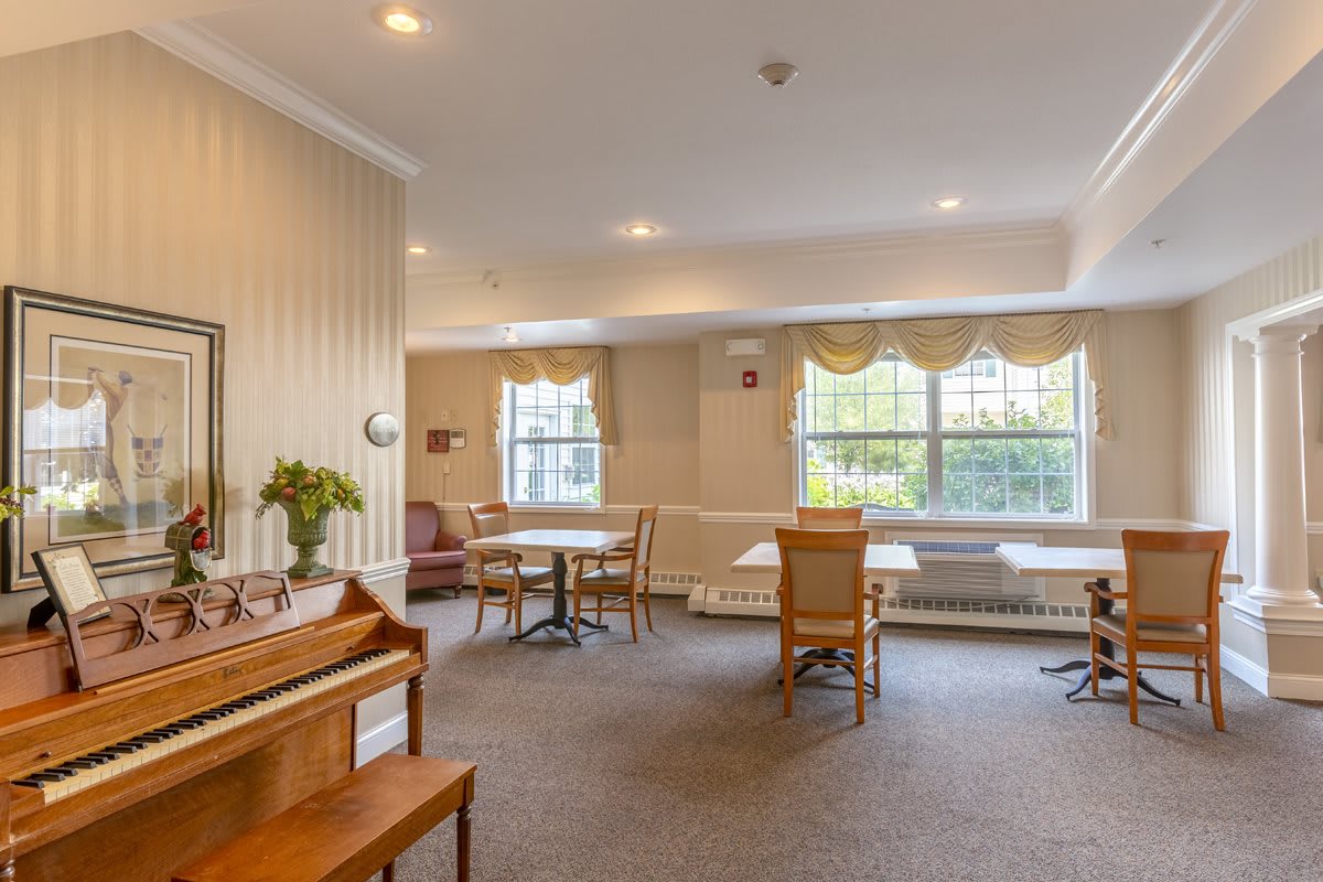 A cozy room interior with tables, chairs, and a piano near large windows.