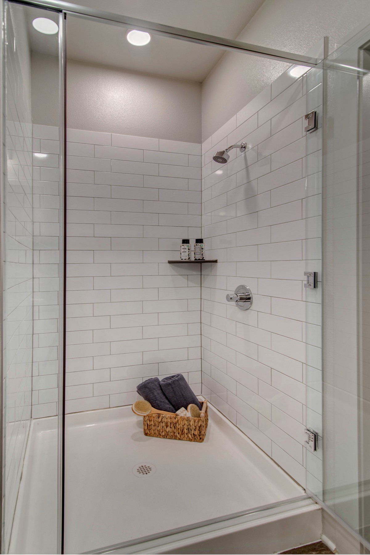 A modern shower with glass doors and white tiled walls, featuring a basket with towels.