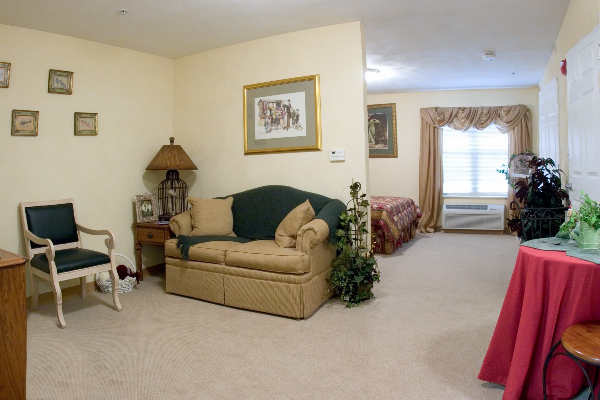 A cozy living room with a sofa and a lamp, leading into a bedroom with natural light.