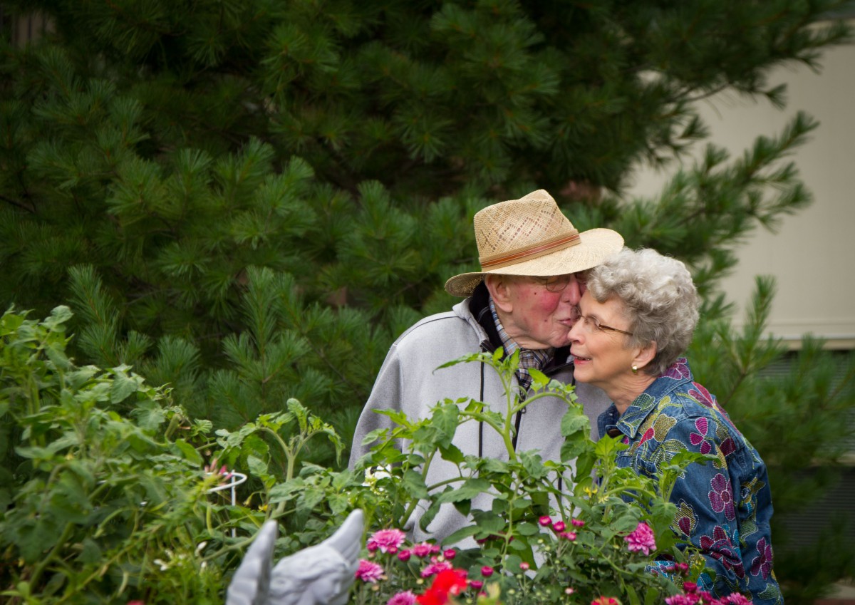 An elderly couple shares a moment of affection while tending to their garden filled with vibrant flowers.