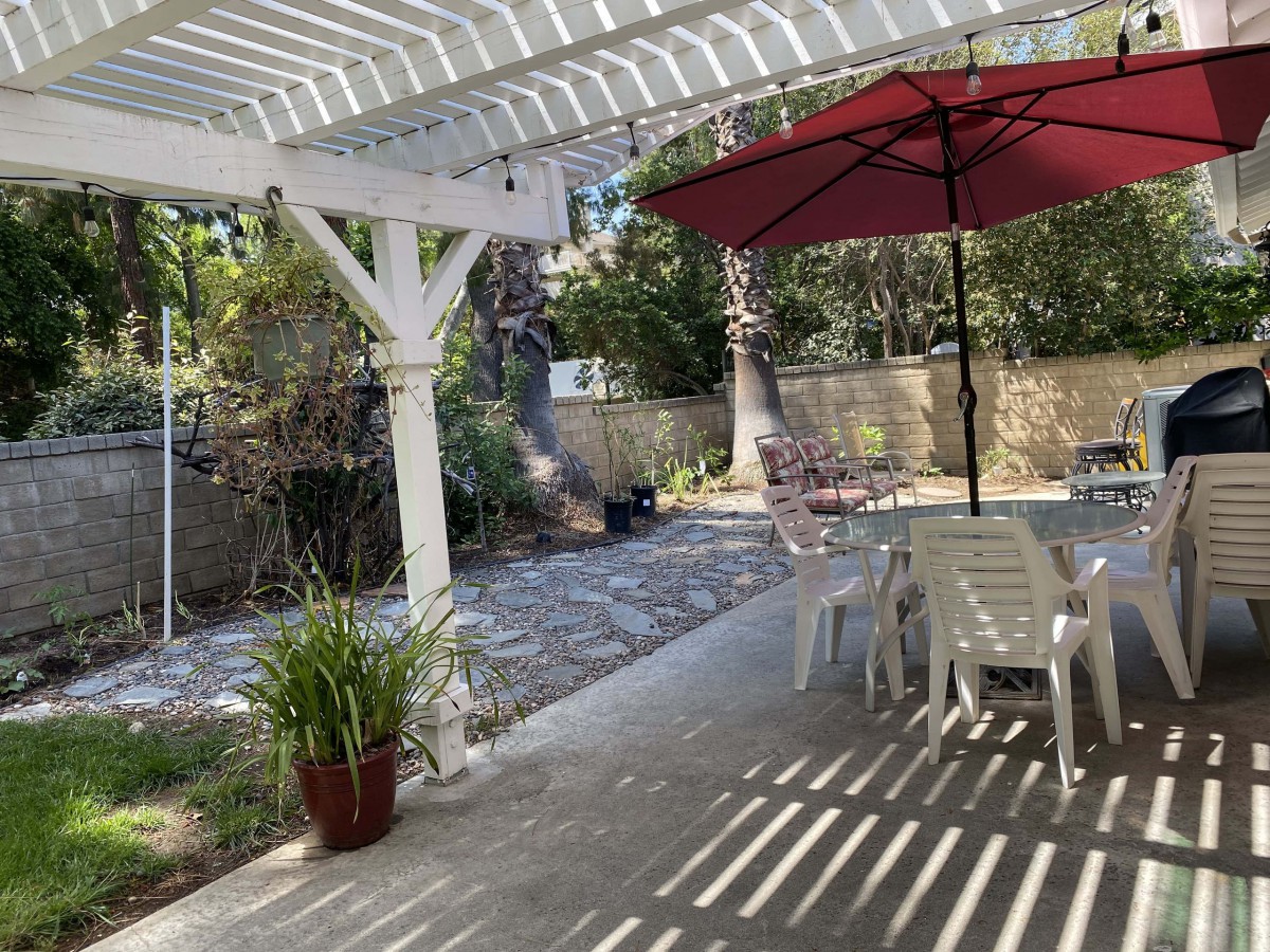A sunny backyard patio with a pergola, an umbrella-covered table, and surrounding greenery.