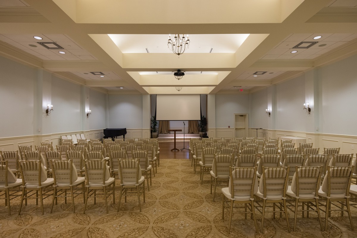 An empty conference room with rows of chairs facing a stage and projector screen.