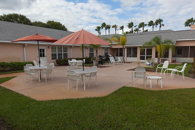 Outdoor patio area with tables, chairs, and umbrellas next to a building surrounded by palm trees.