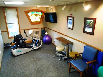 A small exercise room featuring a stationary bike and an exercise ball, with a television mounted on the wall and seating available.