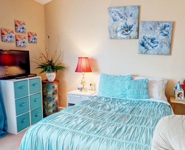 A cozy bedroom featuring a blue color scheme with decorative pillows, artwork, and a bedside lamp.