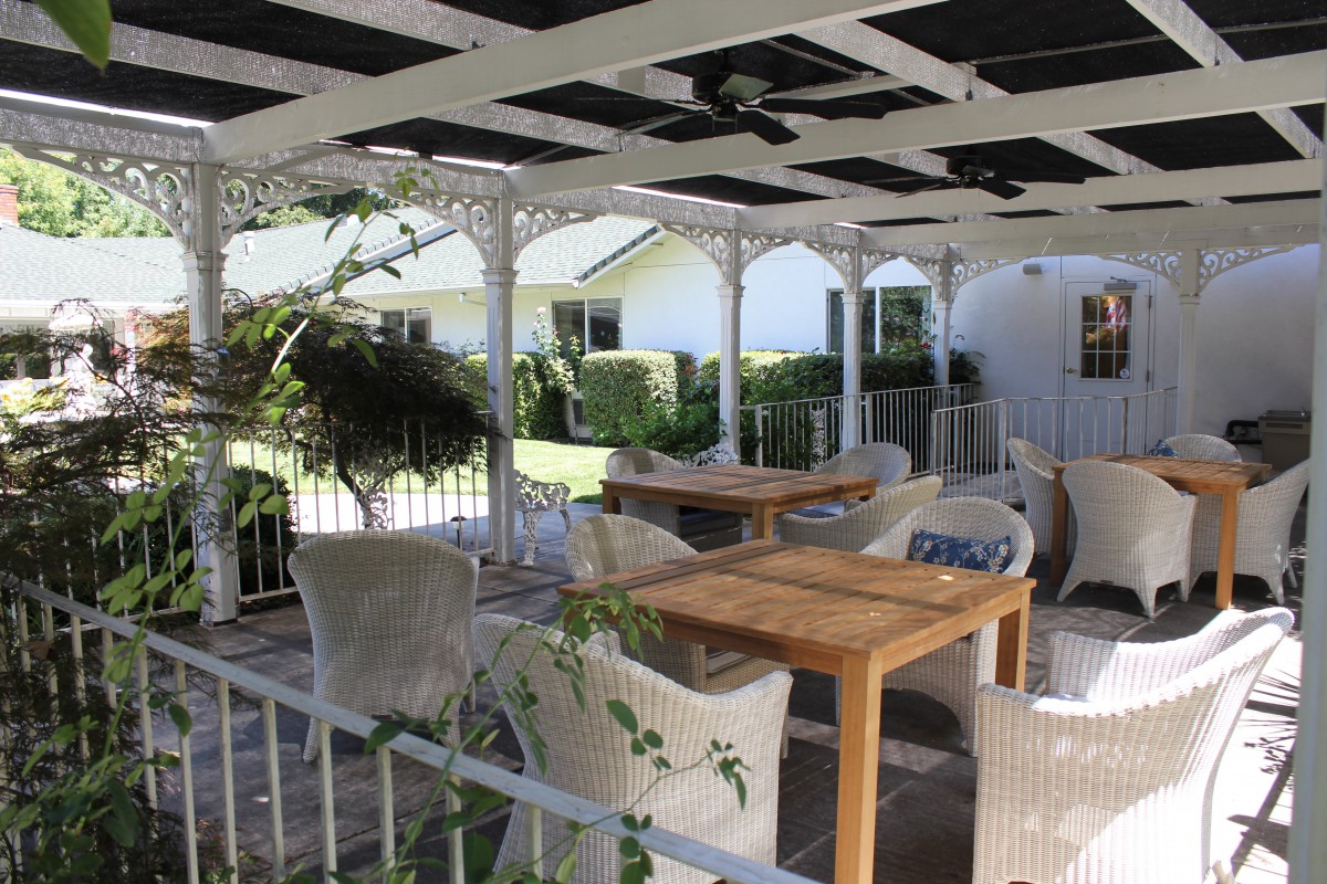 A cozy outdoor patio area with wicker chairs and wooden tables under a pergola surrounded by greenery.
