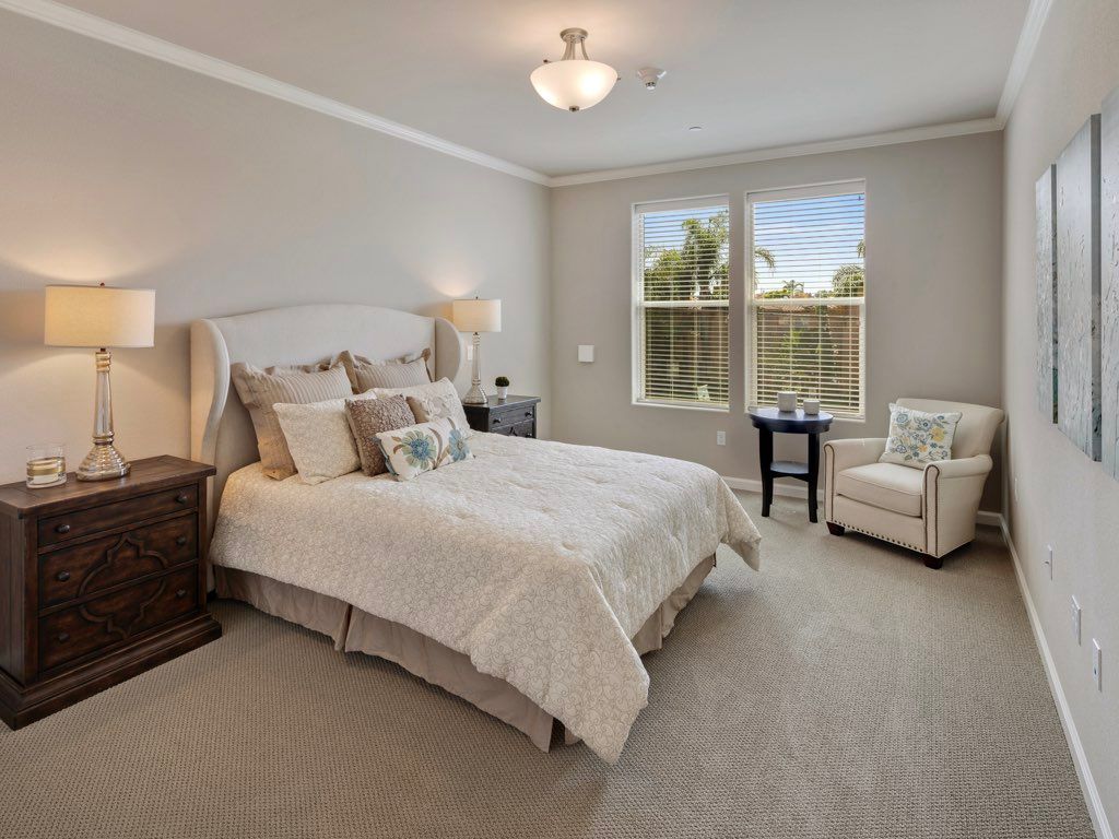 A cozy and elegantly decorated bedroom with neutral tones, featuring a bed, nightstands, and an armchair by the window.