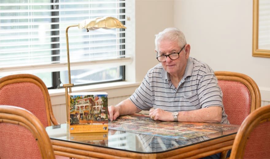 An elderly man wearing glasses is sitting at a desk, reading under a lamp.