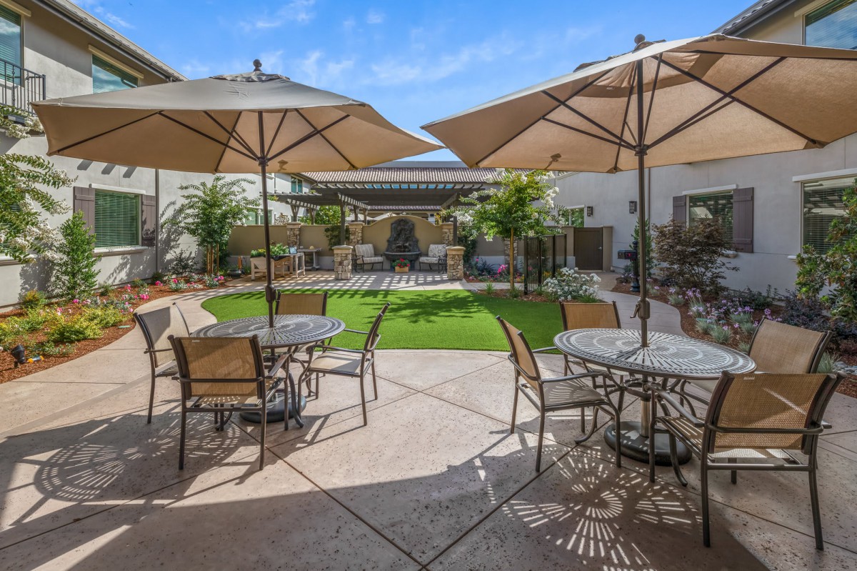 A sunny outdoor patio area with tables, chairs, and large umbrellas providing shade, surrounded by greenery.