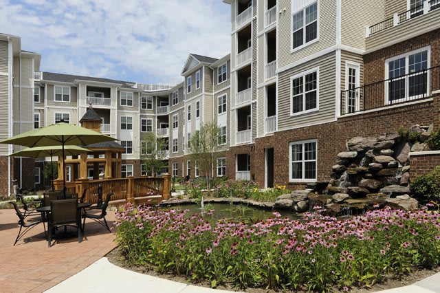 A modern apartment complex with a landscaped garden and pond, featuring outdoor seating areas.