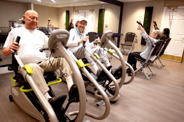 Elderly individuals exercising on stationary bikes in a gym setting.