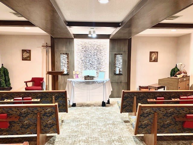An interior view of a small chapel with pews facing the altar adorned with a cross and stained glass windows.