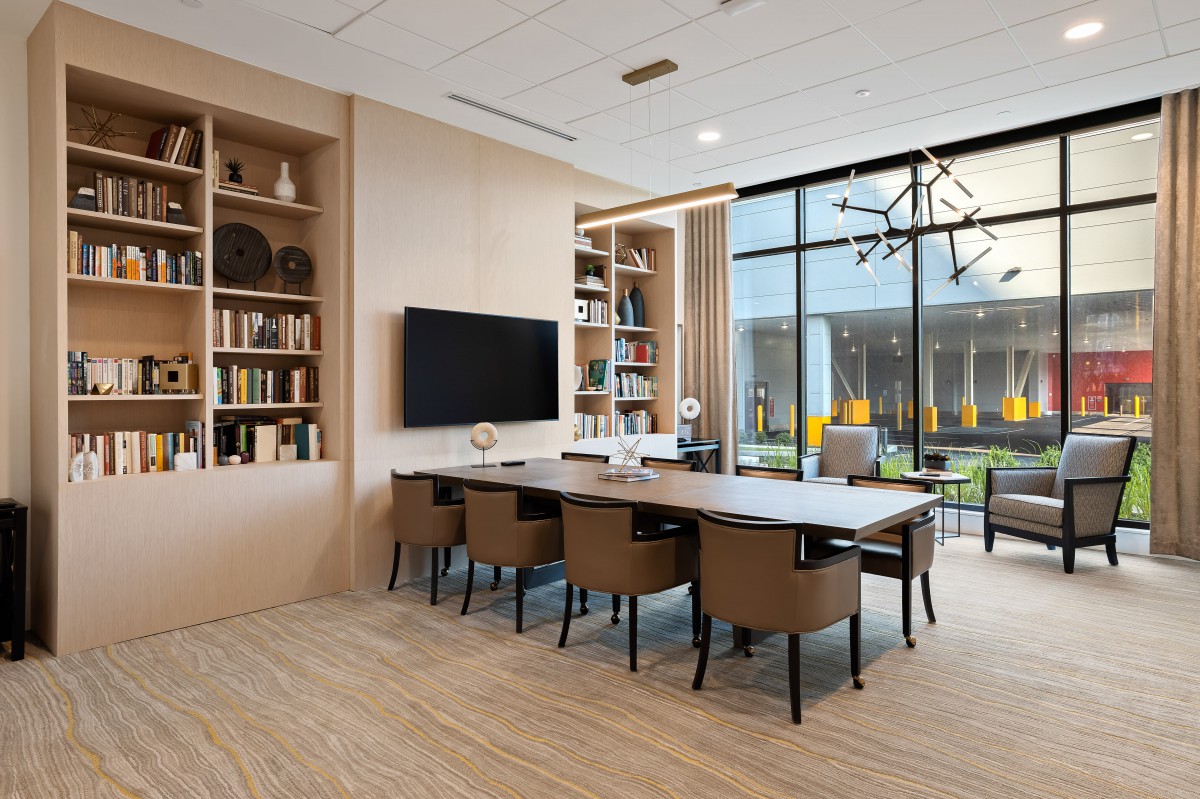 A modern conference room with a large table, chairs, bookshelves, and a television, illuminated by natural light through large windows.