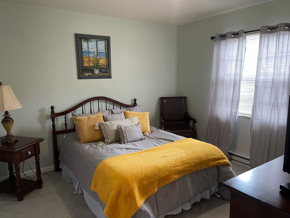 A cozy bedroom with a neatly made bed featuring a yellow blanket and several pillows, complemented by soft lighting from a window.