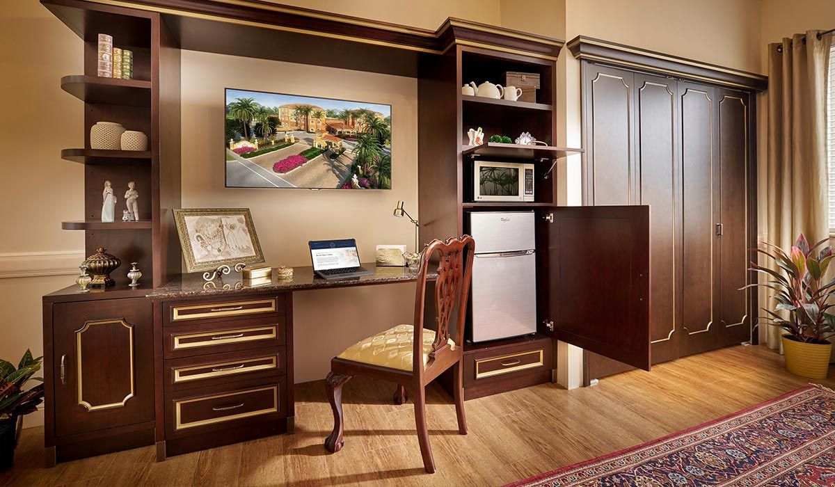 A cozy home office setup with wooden furniture, featuring a desk, chair, laptop, shelving with a microwave and small refrigerator.