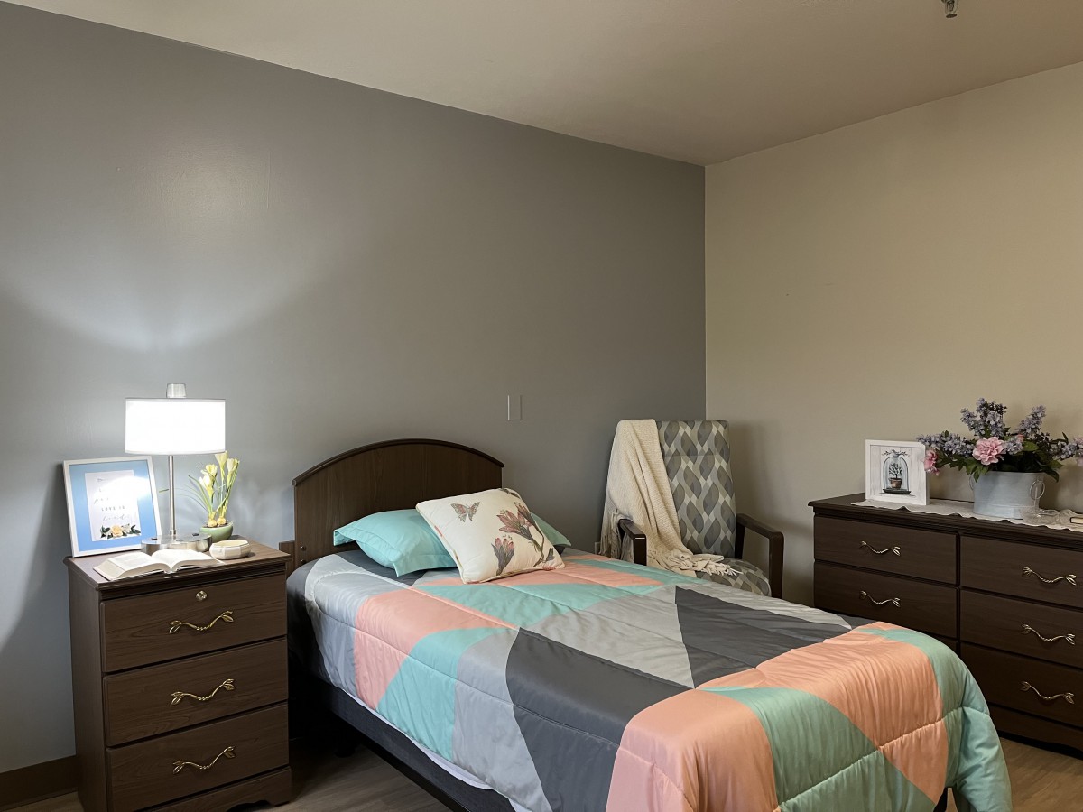 A neatly arranged bedroom with a twin bed, colorful quilt, lamp, and dresser.