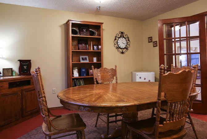A cozy dining room with a wooden table and chairs, a bookshelf in the corner, and a wall clock.