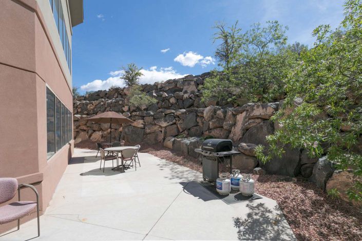 A sunny backyard patio with a table, chairs, and a grill set against a rocky landscaped backdrop.