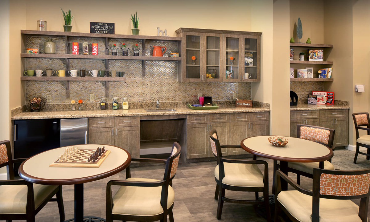 A cozy kitchen interior featuring wooden cabinetry, open shelves with decorative items, and a seating area with tables and chairs.