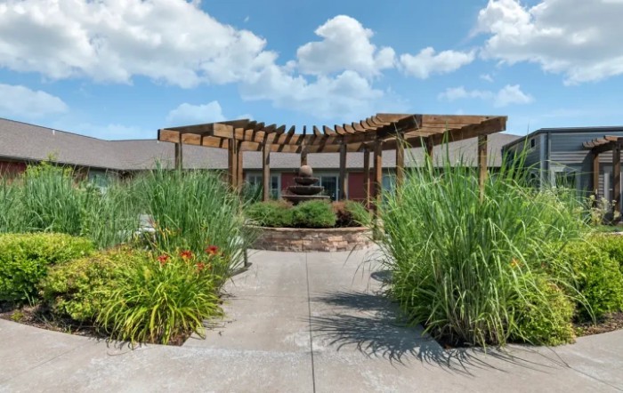 A scenic pathway leads to a wooden pergola surrounded by lush greenery under a bright blue sky.