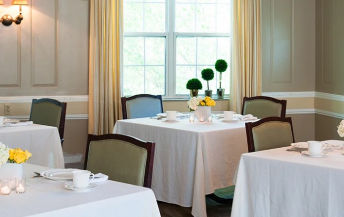 A neatly arranged dining area with tables covered in white tablecloths, decorated with plants and set with tableware near large windows.