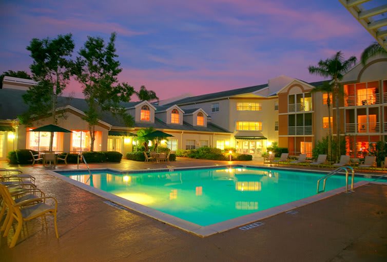 A serene evening view of a hotel with a glowing swimming pool surrounded by illuminated residential buildings.