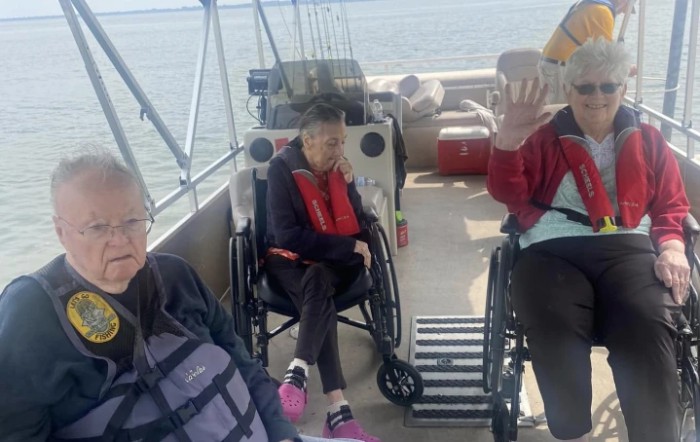 A group of elderly individuals in wheelchairs enjoying a boat ride on a lake, surrounded by fishing rods and wearing life jackets.
