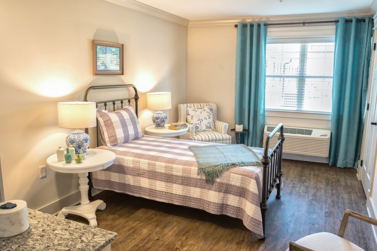 A cozy bedroom with a neatly made bed, soft lighting, and comfortable seating by the window.
