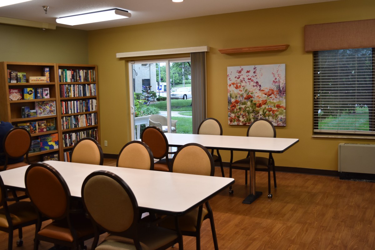A library interior featuring tables and chairs, bookshelves, artwork, and a window with a view outside.