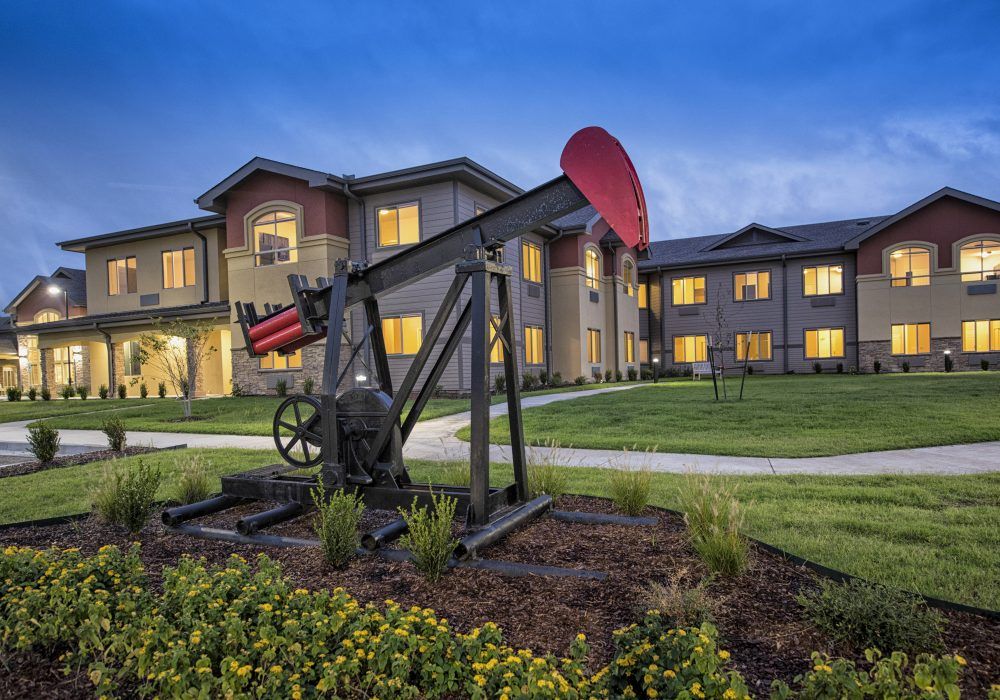 An oil pumpjack in front of a lit residential building during the evening.