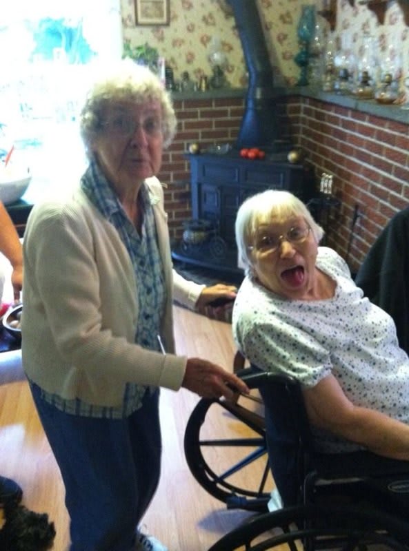 Two elderly women sharing a moment indoors, one seated in a wheelchair.