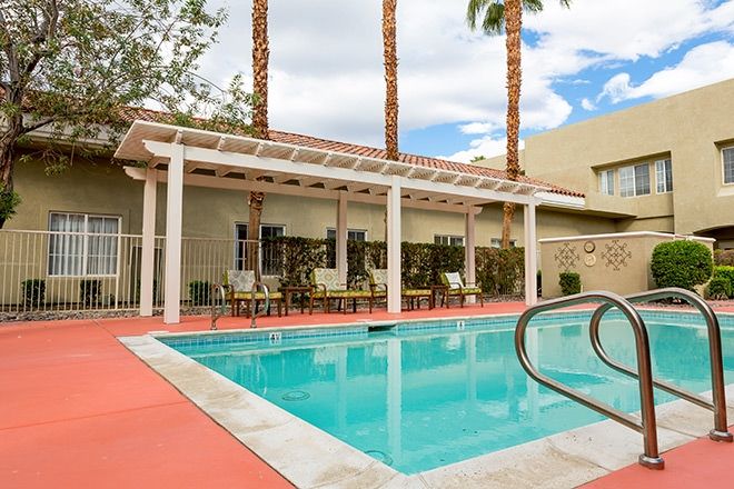 An outdoor pool area with seating under a pergola and palm trees in the background.