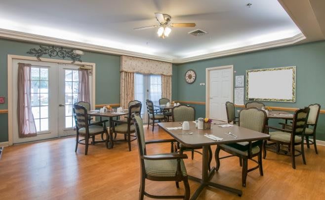 A dining room with multiple tables and chairs arranged on a hardwood floor, featuring a ceiling fan and large windows with curtains.