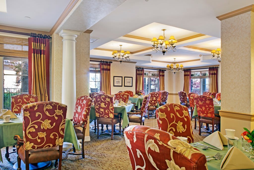 An elegant dining area with upholstered chairs and chandeliers, featuring tables covered with tablecloths.