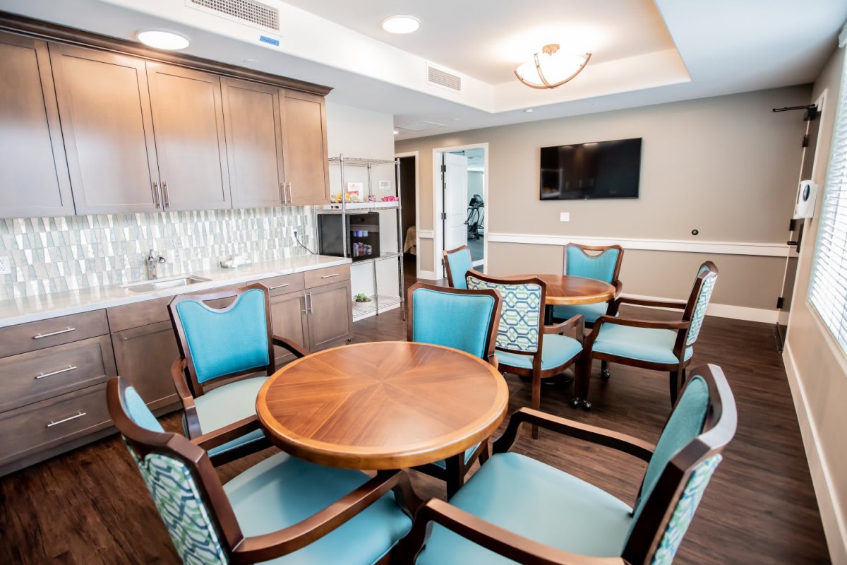 A modern dining area with wooden tables and blue upholstered chairs, featuring a kitchenette and a wall-mounted television.