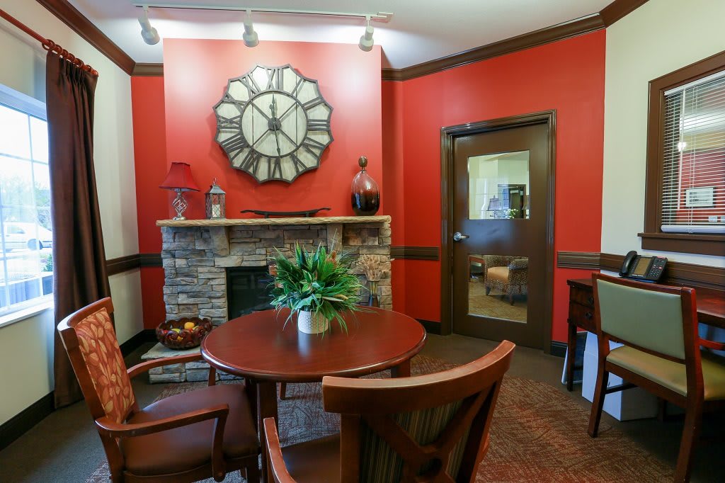 A cozy office meeting room with red walls, featuring a round table, chairs, a large decorative clock above a stone fireplace, and a potted plant.