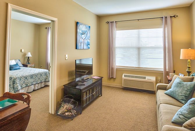 A cozy living room with a TV stand, sofa, and a view into the adjacent bedroom. The space is softly lit through a large window with curtains.