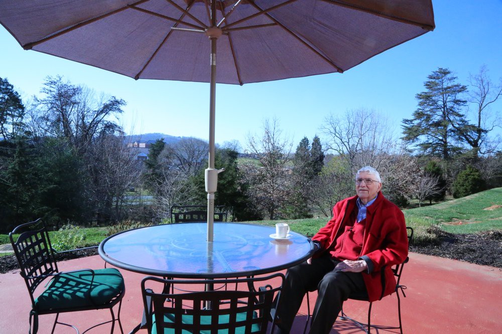 An elderly person sitting at a patio table under an umbrella, enjoying a sunny day with a scenic view in the background.