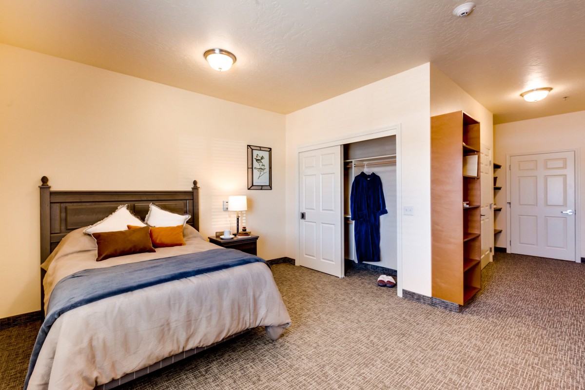 A cozy bedroom with a neatly made bed, open closet, and bedside table with a lamp.