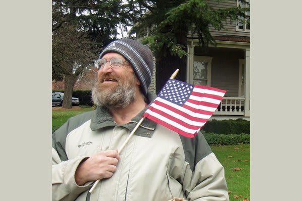 A bearded man wearing a beanie and jacket is holding an American flag while standing outdoors in front of a house.