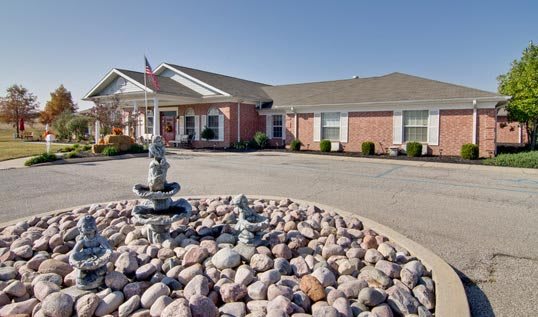 A single-story brick building with a circular driveway featuring a fountain surrounded by rocks.