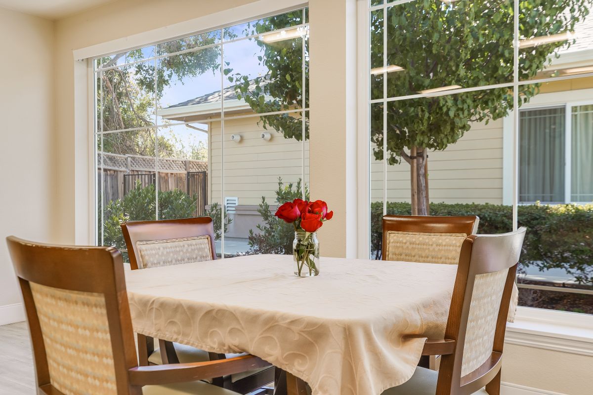 A cozy dining area with a table set for four and a vase of red flowers, surrounded by large windows overlooking a garden.