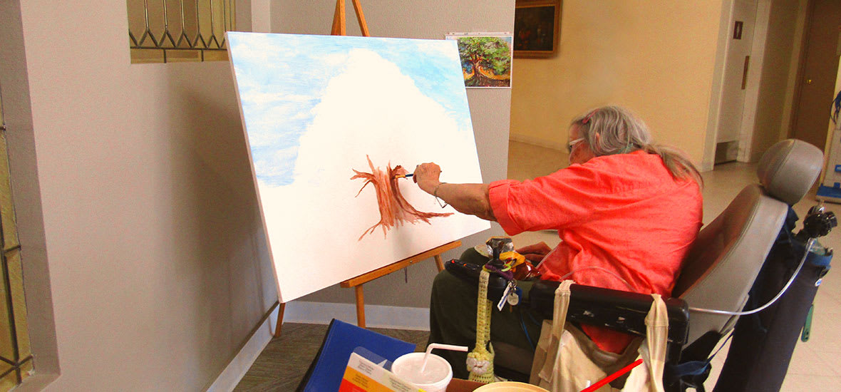 An artist in a wheelchair working on a painting with vibrant colors on a canvas inside a studio.