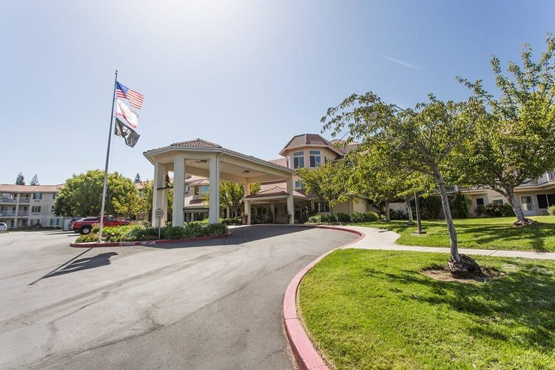 A well-maintained building entrance with a circular driveway and flags, surrounded by lush trees and landscaping on a sunny day.