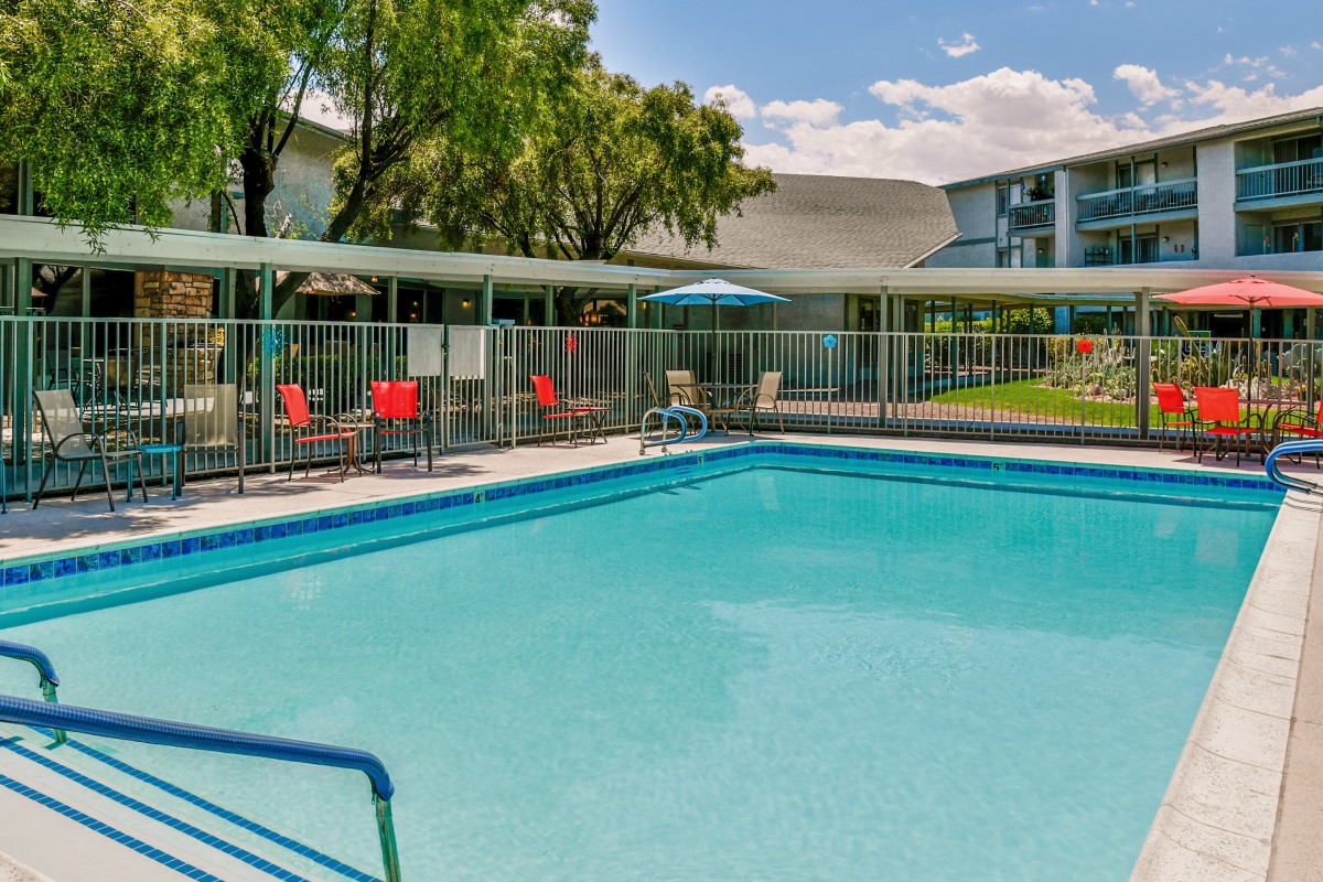 An outdoor swimming pool surrounded by a fence, with chairs and trees in the background.