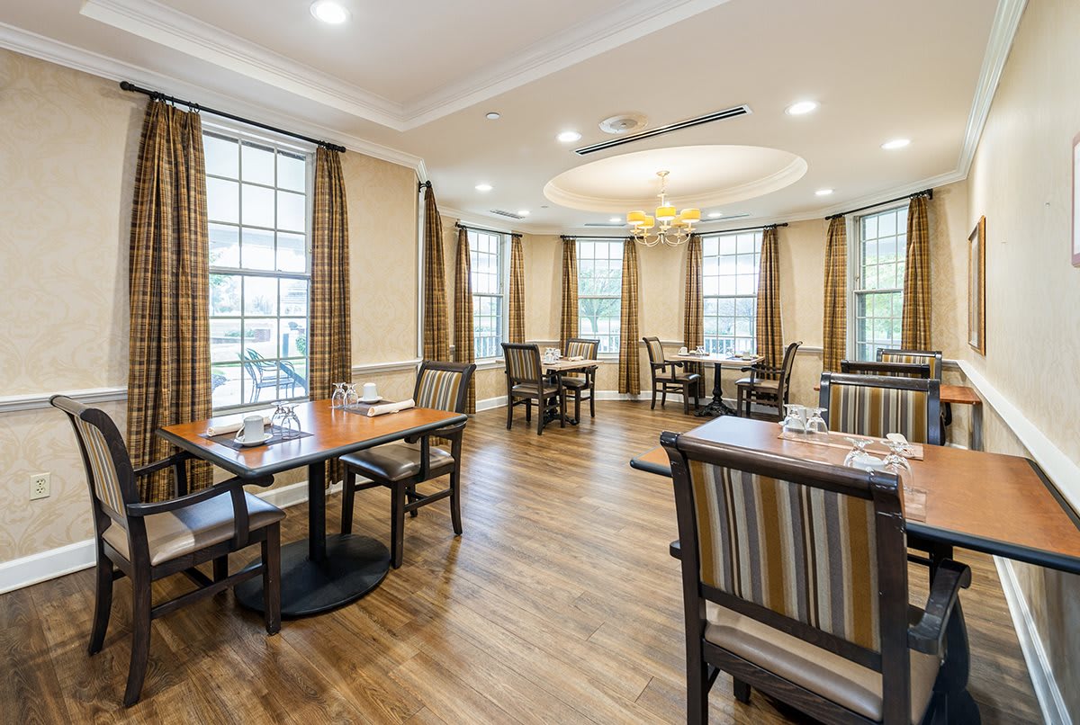 A cozy dining area with neatly arranged tables and chairs, featuring large windows and a central chandelier.