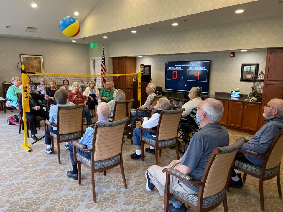 A group of elderly individuals enjoying a social activity indoors, seated in a circle and playing with a beach ball.