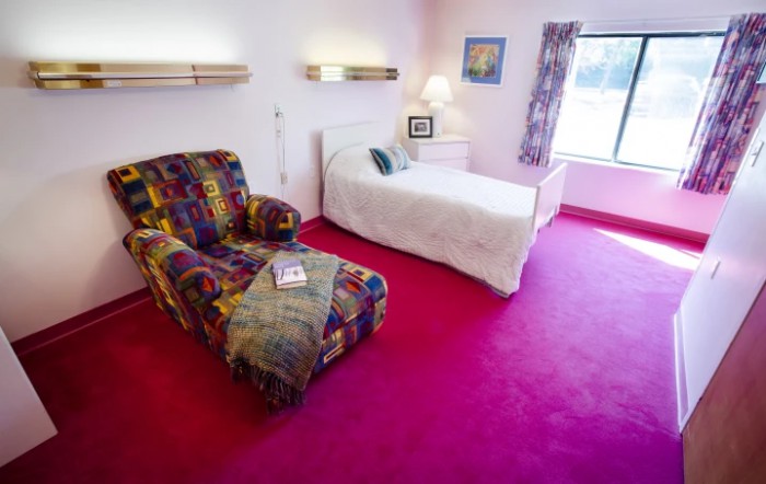 A cozy bedroom with a vibrant pink carpet, featuring a single bed, a colorful armchair, and a window letting in natural light.