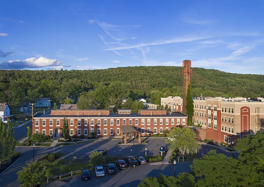 A historic brick factory building with a tall chimney, surrounded by greenery and a clear blue sky.